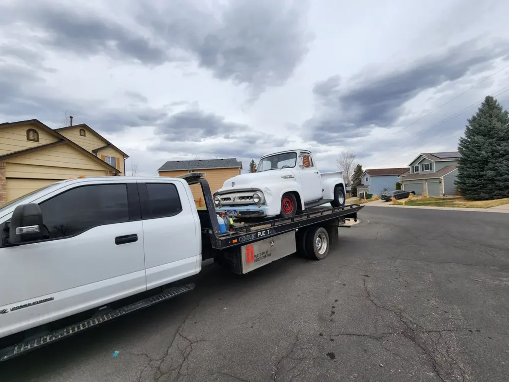 Flatbed carrying an old white classic pickup truck down a neighborhood street, we buy junk cars in the Thornton, CO area
