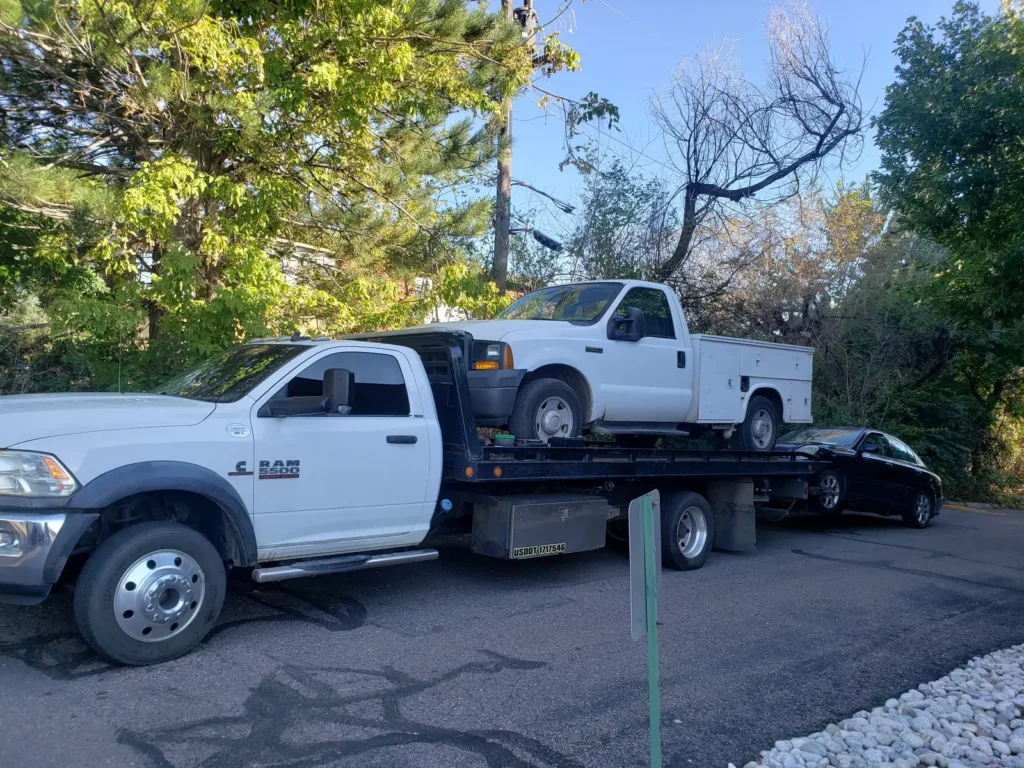 Ram flatbed hauling a white utility work truck with a car in tow, cash for junk cars in the Watkins, CO area