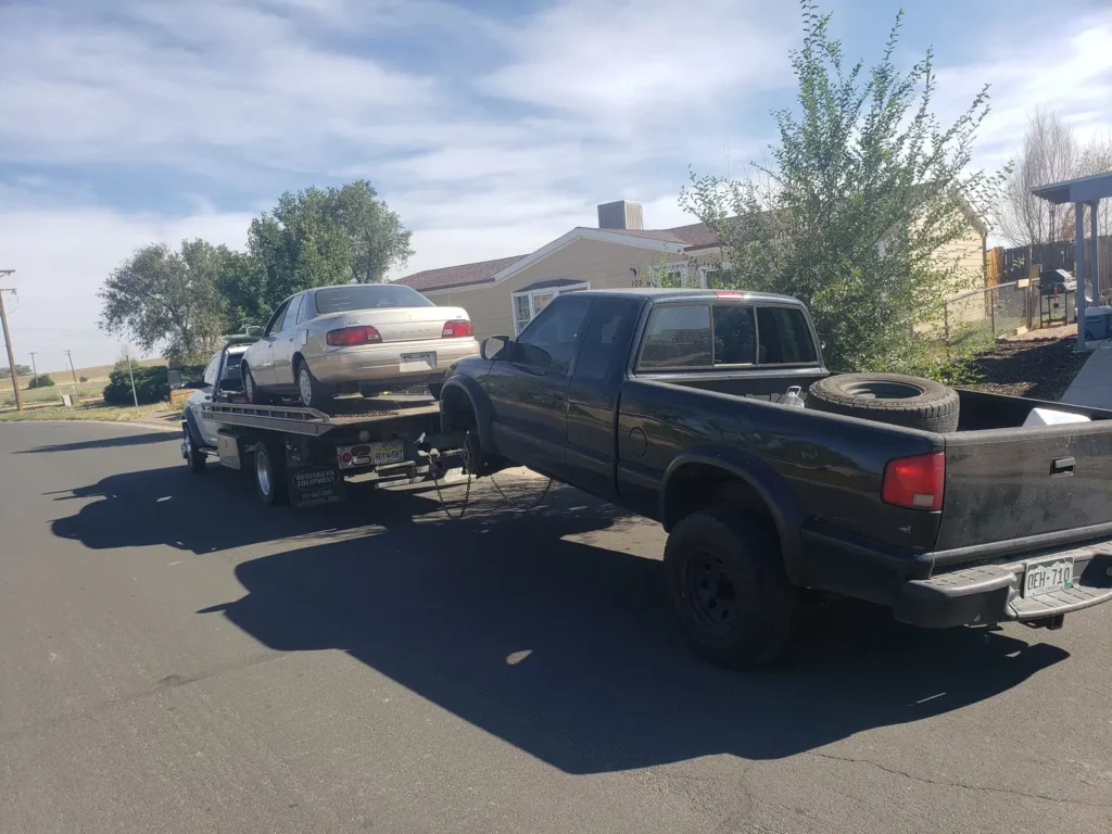 Flatbed hauling a sedan with a black pickup in tow, we buy junk cars in the Loveland, CO area