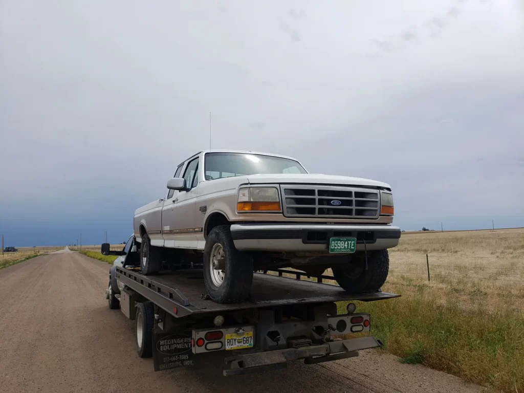 Old white Ford pickup loaded on a flatbed on a rural road, we buy junk cars in the Henderson, CO area